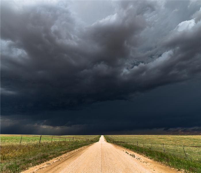 Large, dark clouds overlook a dirt road and open fields.